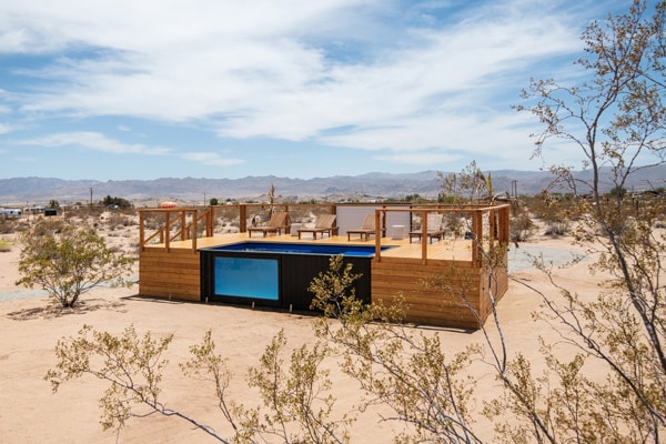shipping container swimming pool with a window on it in the middle of the the Joshua Tree desert with a large wooden deck next to the pool